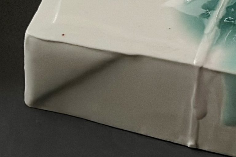 Detail image of a porcelain canvas that looks like a painter’s canvas fabric folding over the edge. Glaze colors are turquoise, dark blue, and light green.